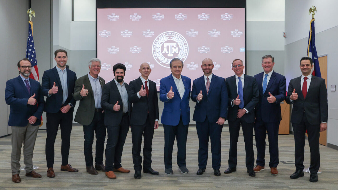 A photo of several men with their thumbs up in front of a Texas A&M Logo projected onto a screen.