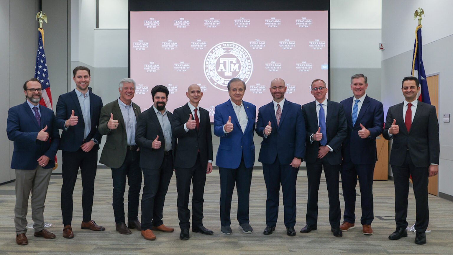 A photo of several men with their thumbs up in front of a Texas A&M Logo projected onto a screen.