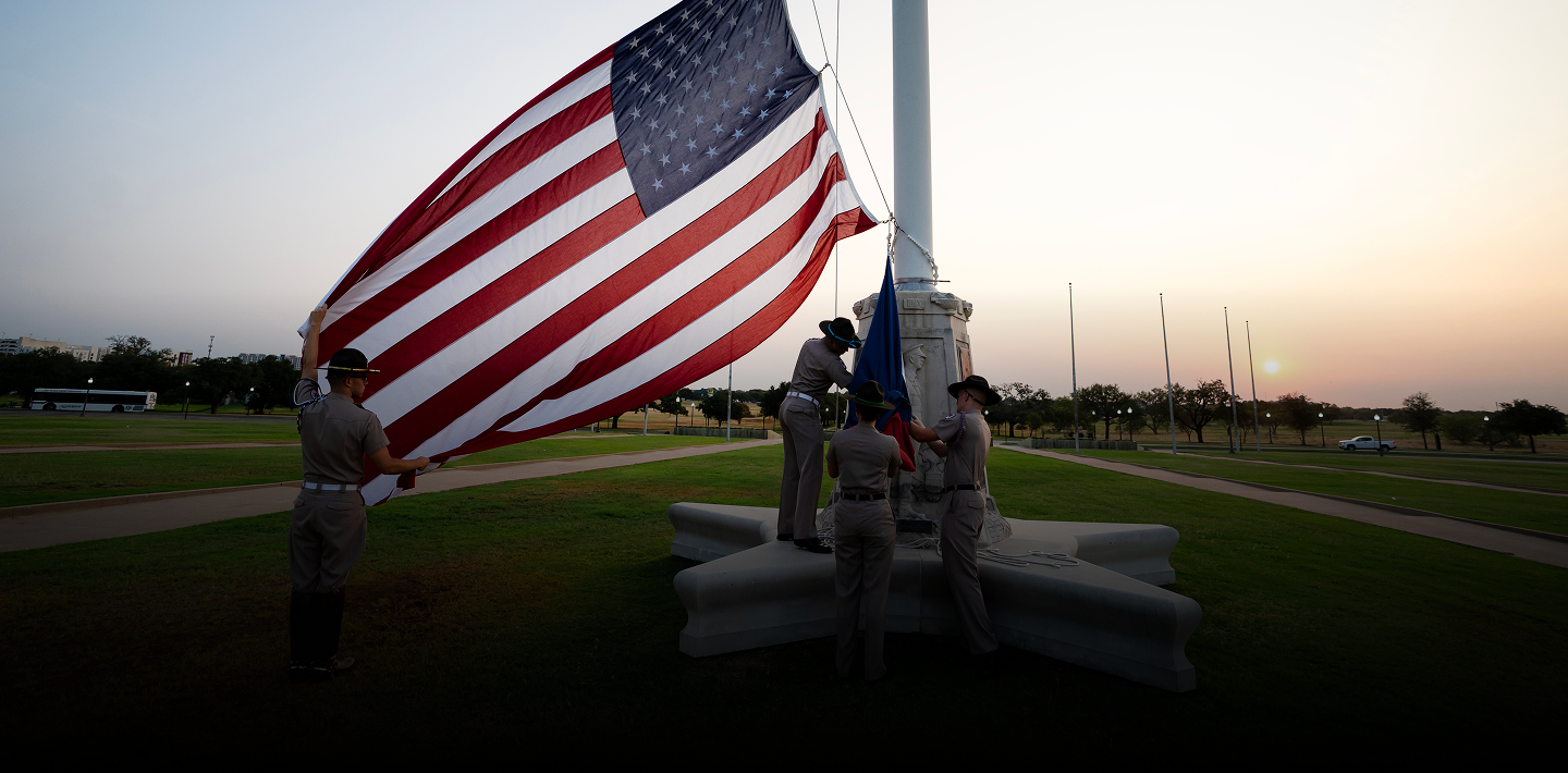Four Texas A&M University Corps of Cadets members in tan uniforms raising an American flag at sunrise.