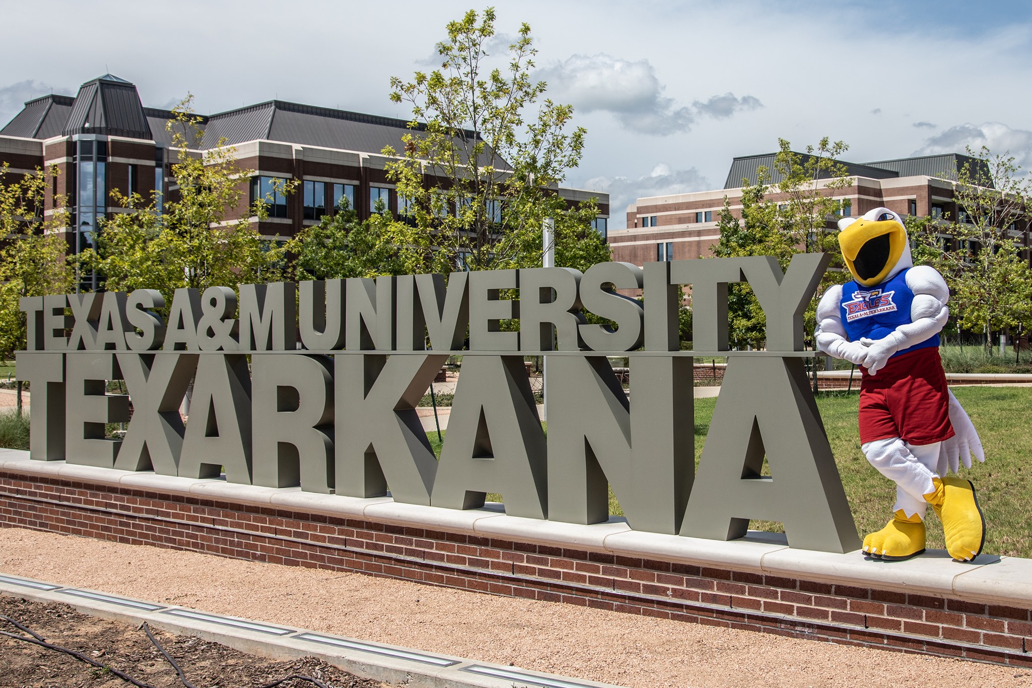 A mascot stands beside a large sign for Texas A&M University-Texarkana, with campus buildings and trees in the background.