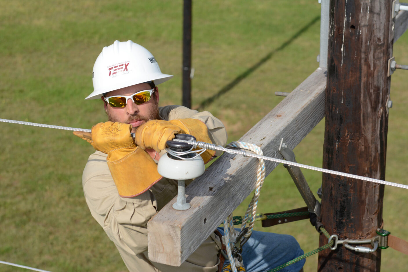 A utility worker wearing a hard hat and gloves is securing a wire to a wooden utility pole.