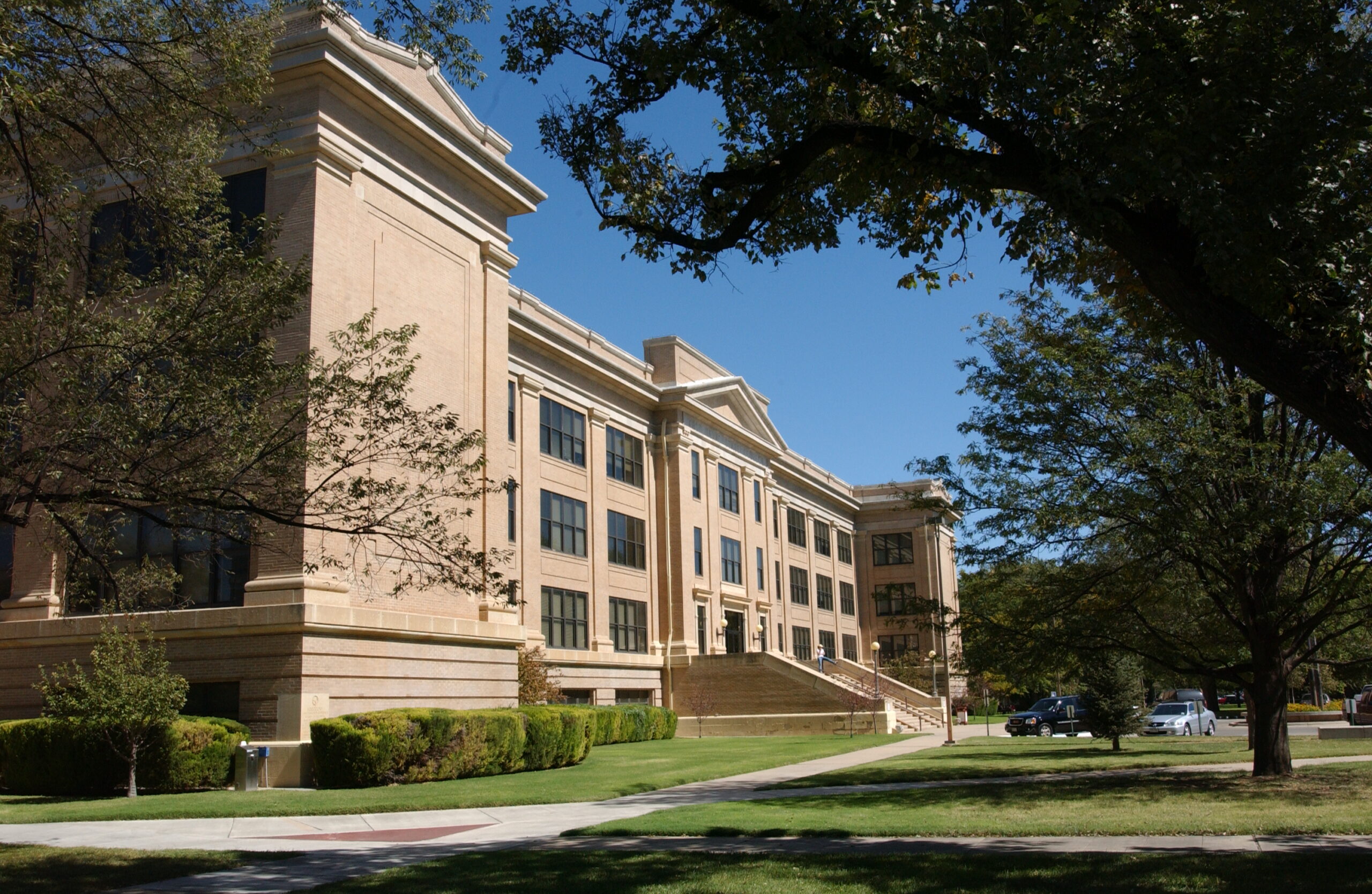 A large, light-colored stone building with classical architectural details stands under a clear blue sky, surrounded by green lawns and trees.