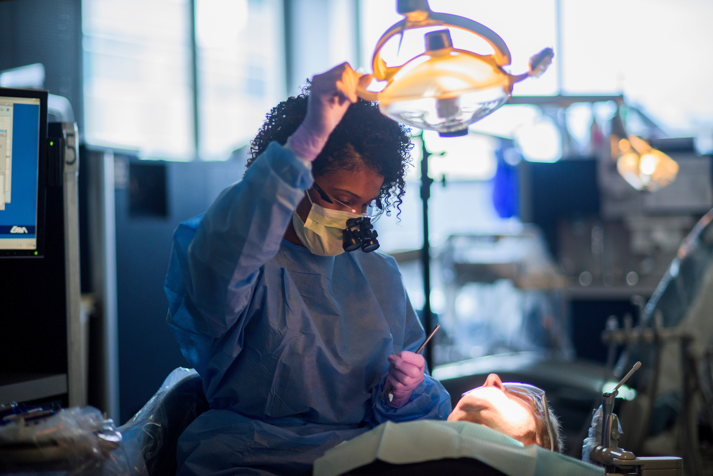 A dental professional in scrubs and a mask adjusts a surgical light above a patient undergoing treatment.
