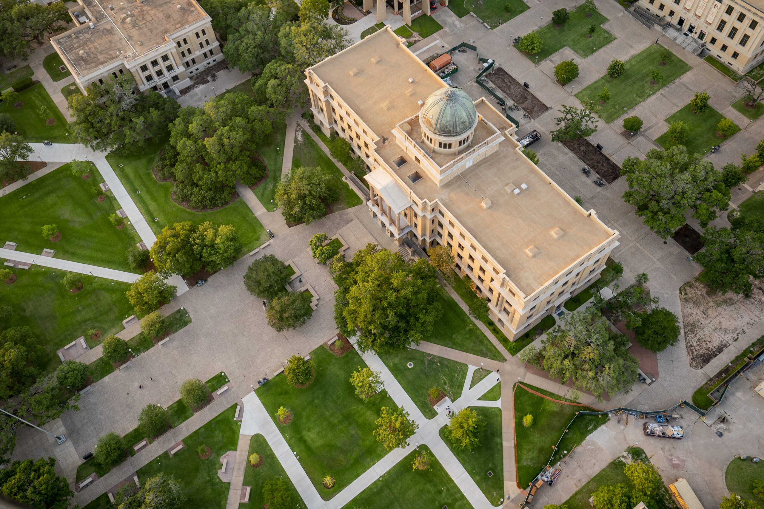 An aerial view of a university campus featuring a prominent building with a dome, surrounded by green lawns, trees, and paved walkways.
