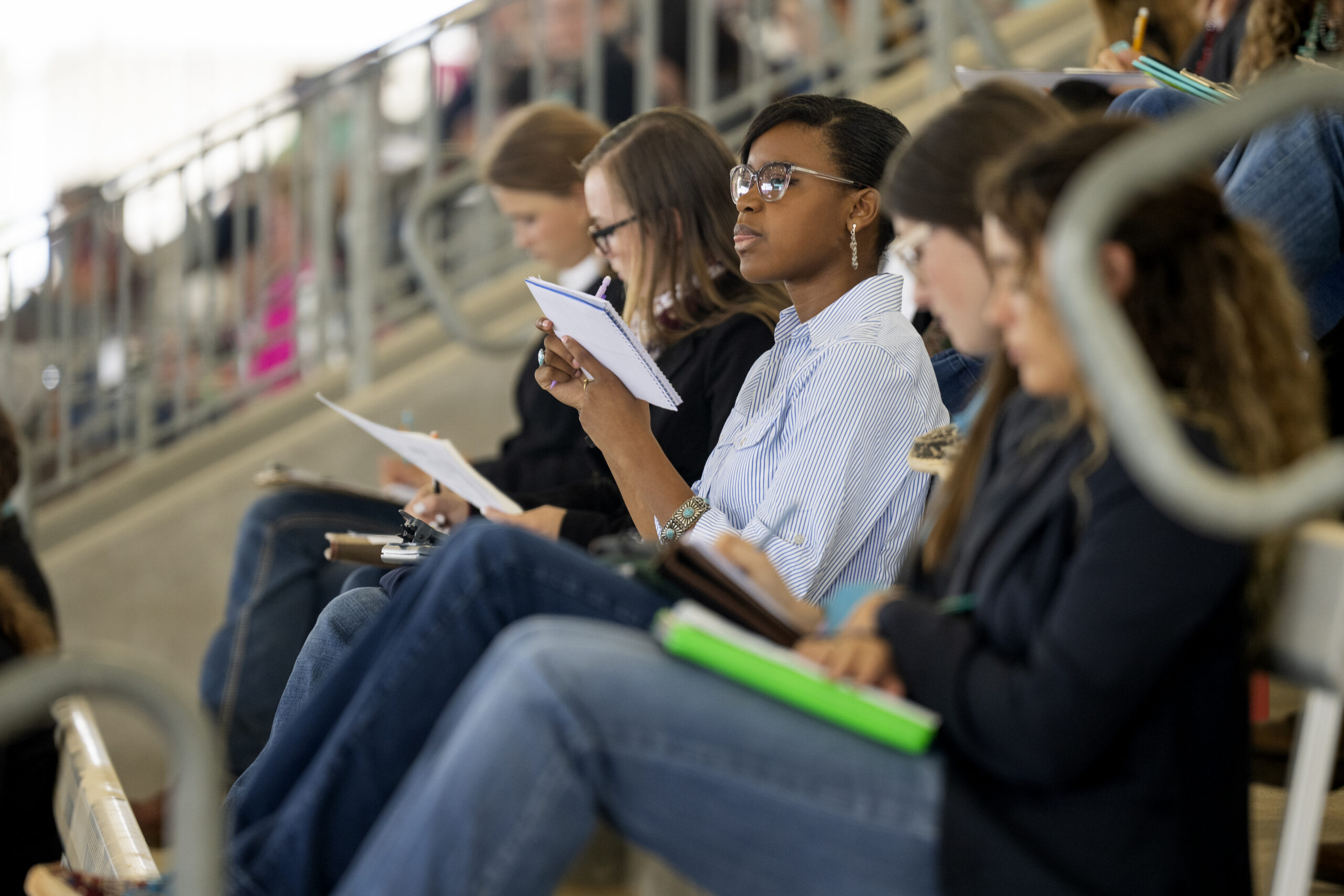 A group of individuals sit in stadium-style seating, attentively taking notes with notebooks and pens.