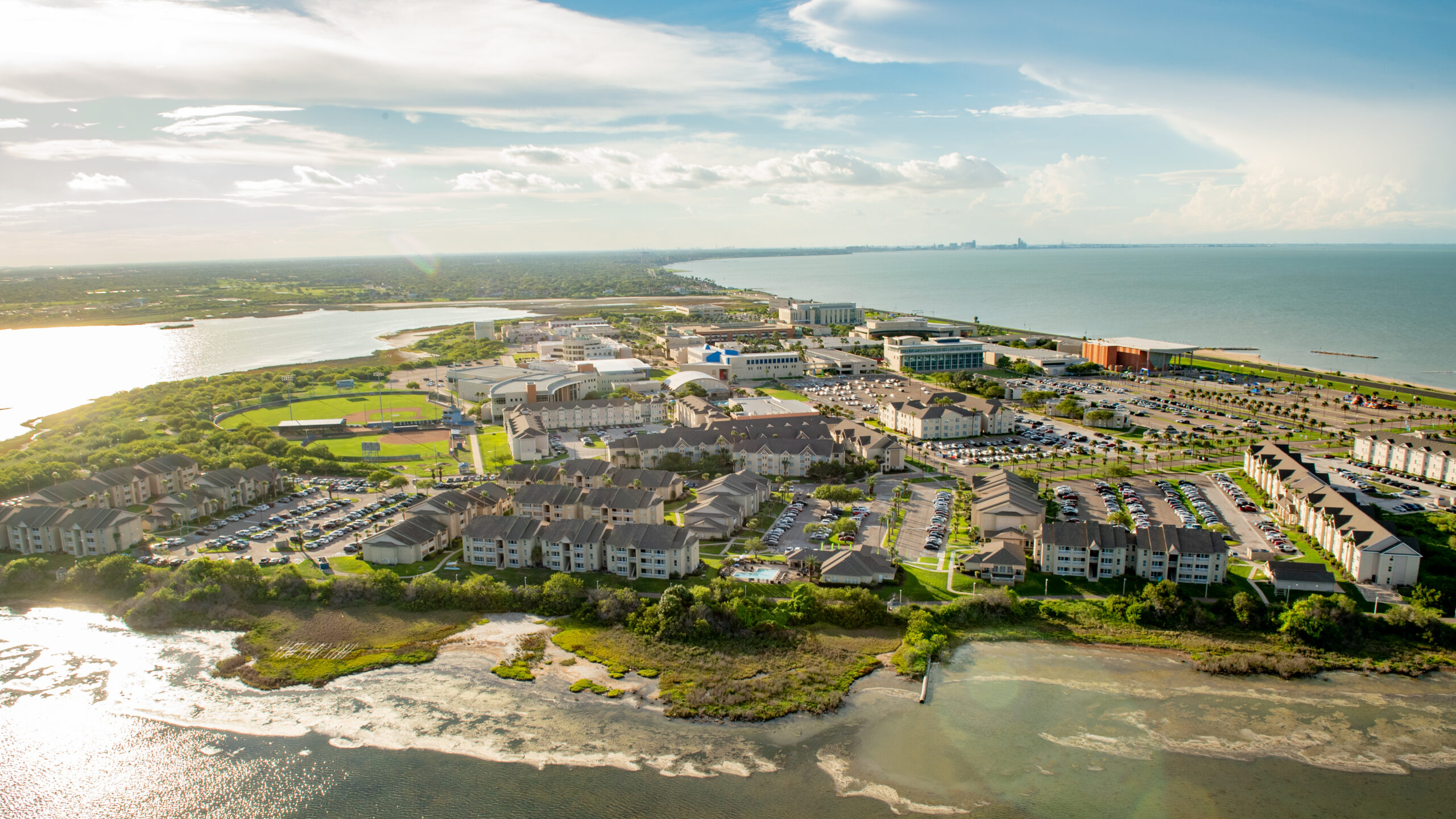 An aerial view of a coastal residential complex with apartment buildings, parking lots, and a sports field, situated between a body of water and a larger sea under a partly cloudy sky.
