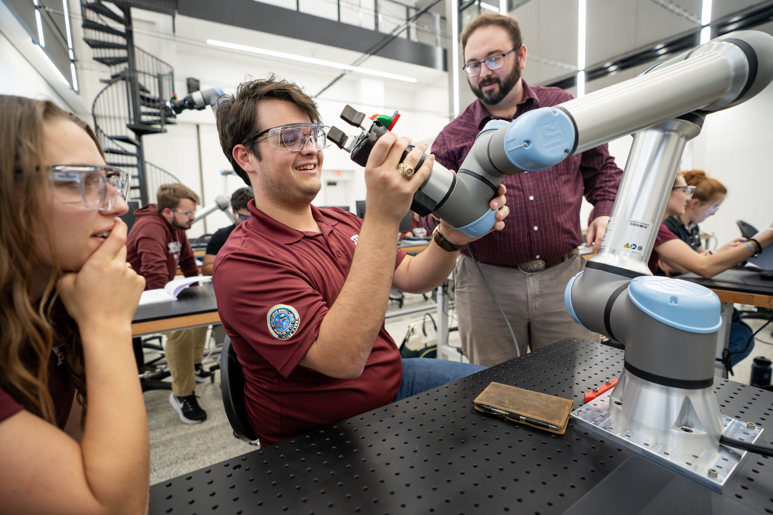 A student operates a robotic arm in a lab setting while peers observe and assist.