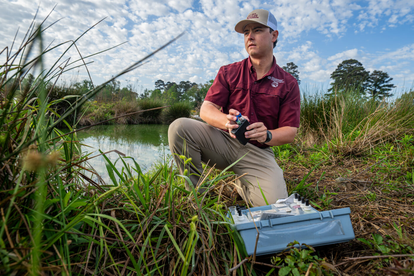A person in a maroon shirt and cap kneels by a pond, holding a small device while a blue case with test tubes rests nearby in tall grass.