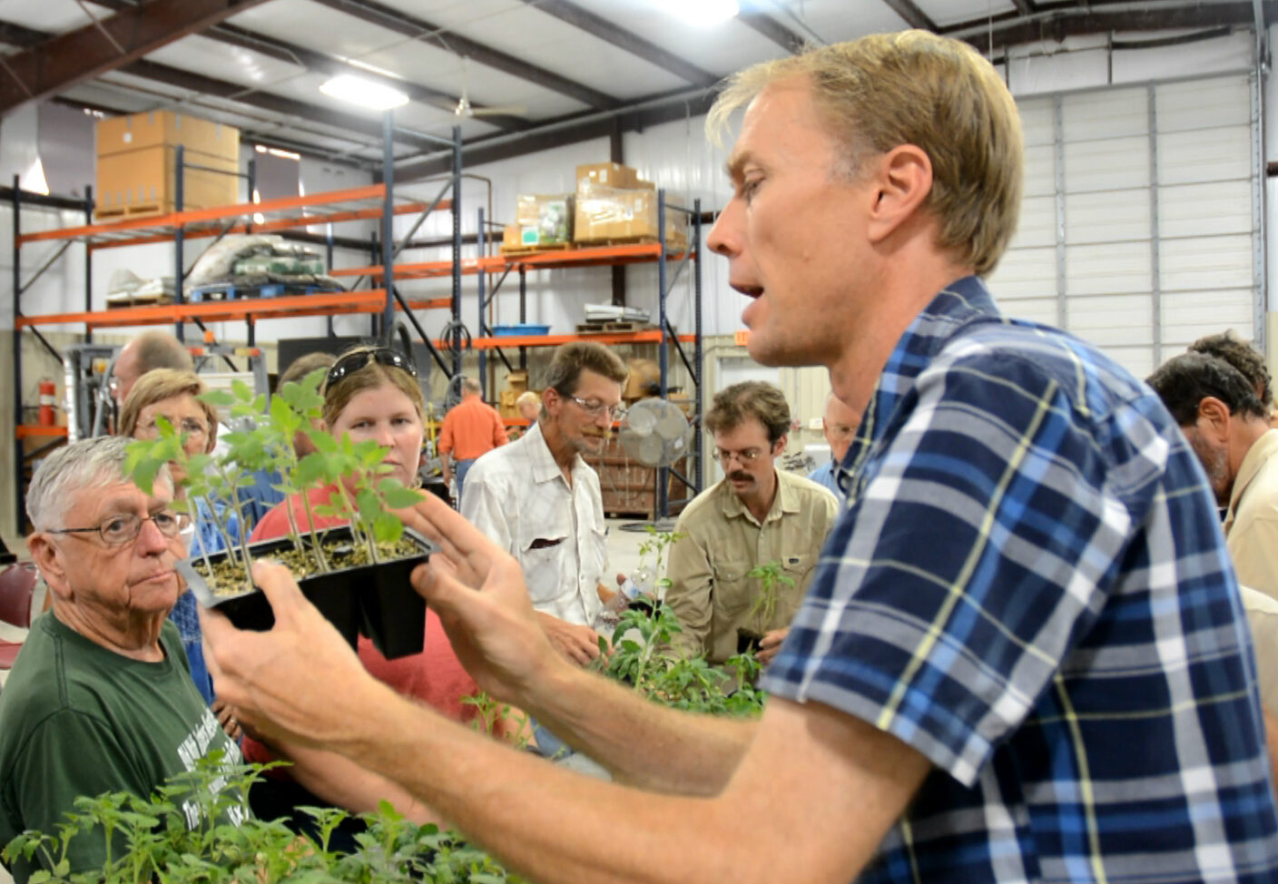 A person in a plaid shirt holds a tray of young plants while speaking to a group of attendees in a warehouse setting.