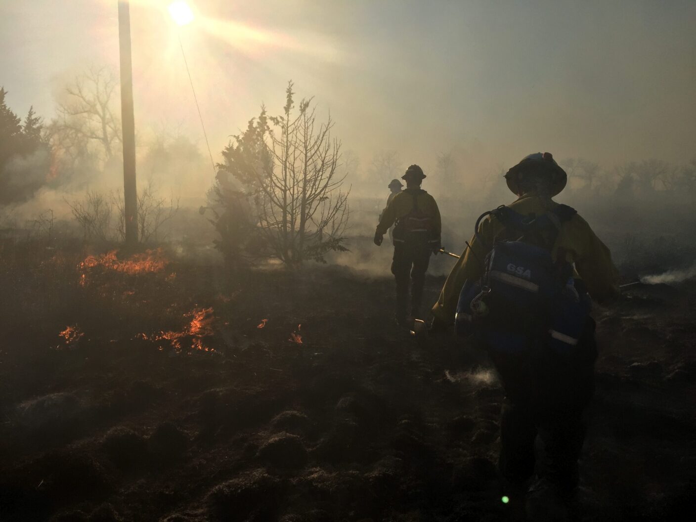 Firefighters navigate through smoke and flames in a wildfire scene, with the sun shining brightly behind them.