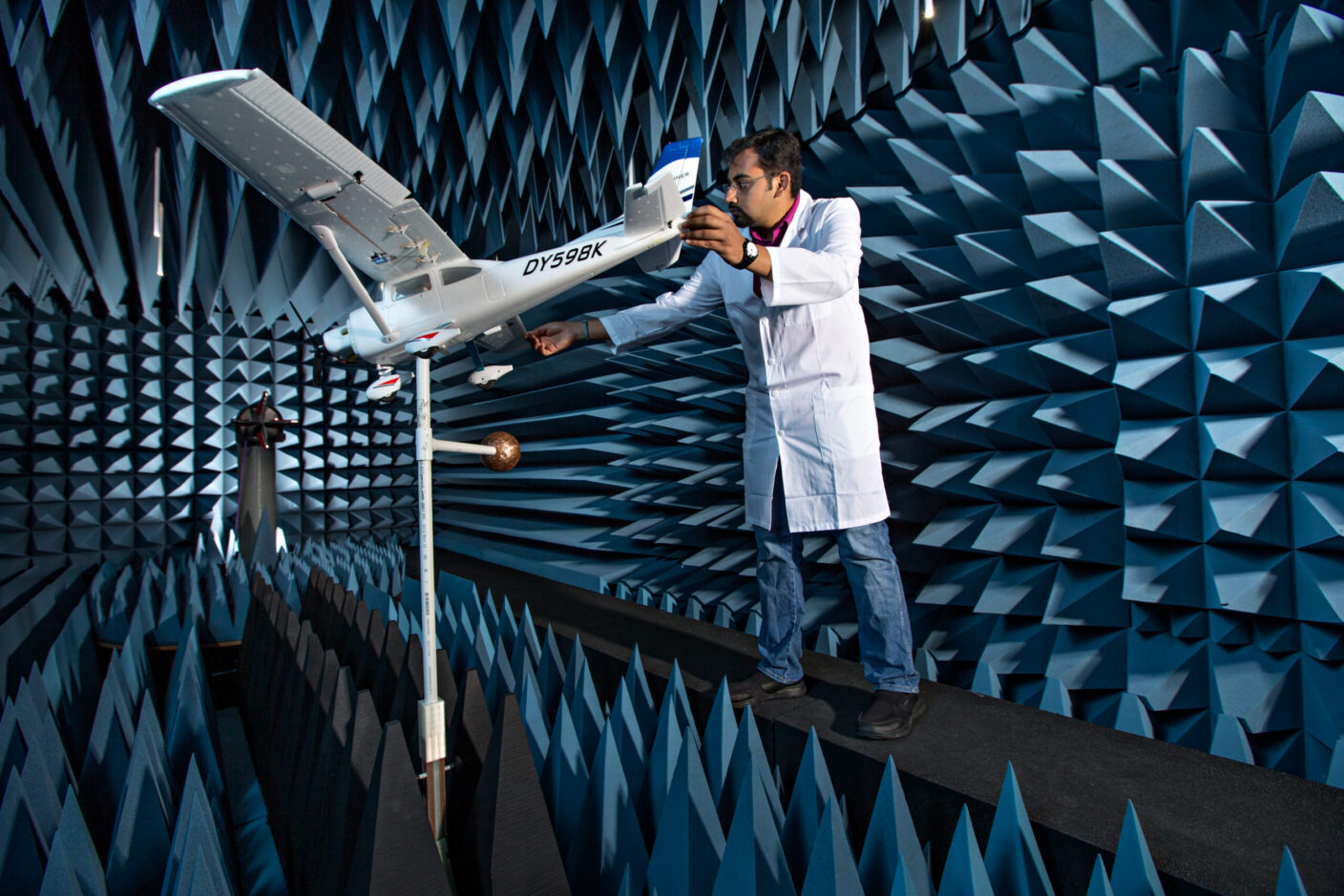A technician in a lab coat uses a device to measure the acoustic properties of a small aircraft inside a soundproof testing chamber.