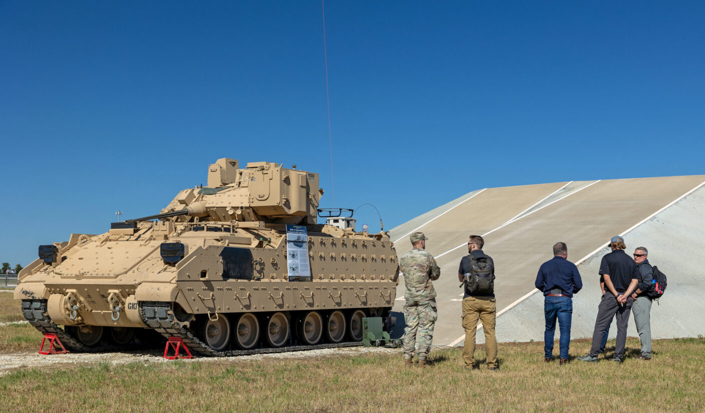 A group of individuals observes a tan armored vehicle on display outdoors under a clear blue sky.