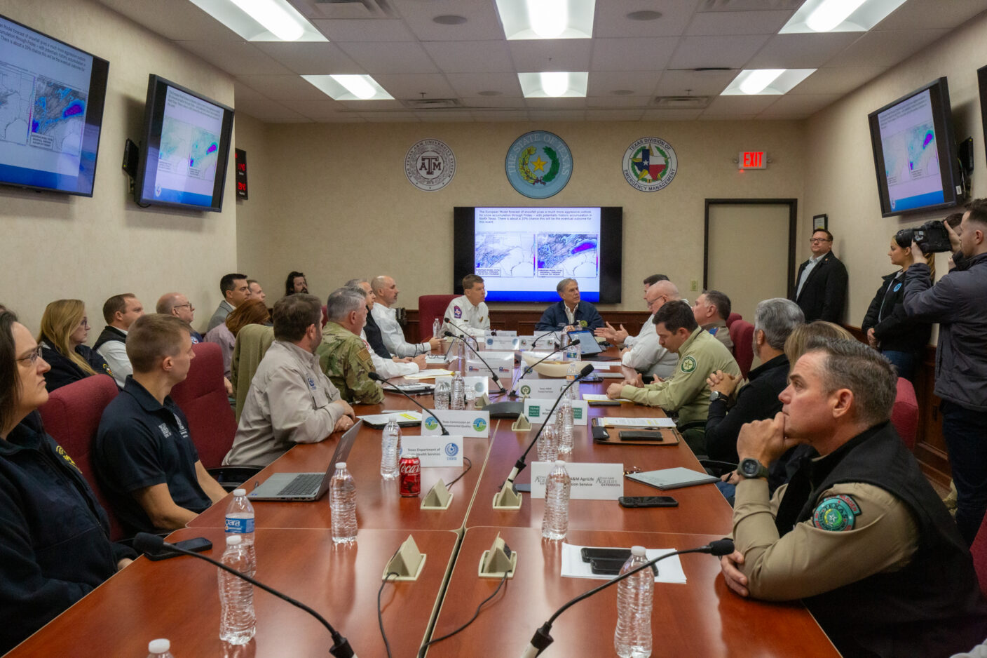 A group of professionals gathers around a long conference table in a meeting room, reviewing maps and data displayed on multiple screens, with some individuals taking notes and others observing attentively.