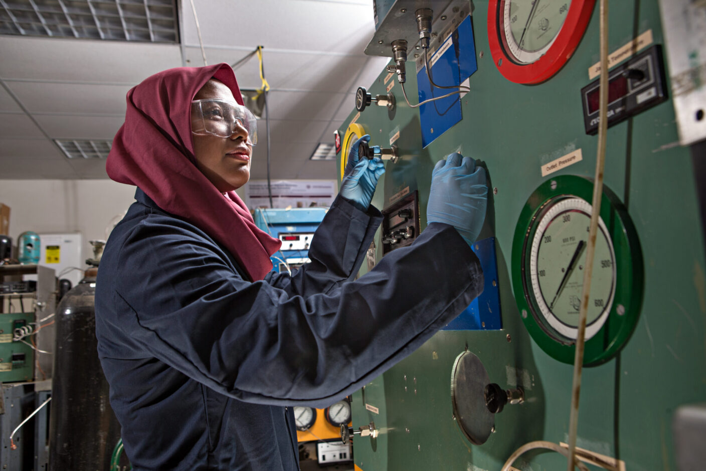 A person in protective gear operates machinery with gauges and valves in a laboratory setting.