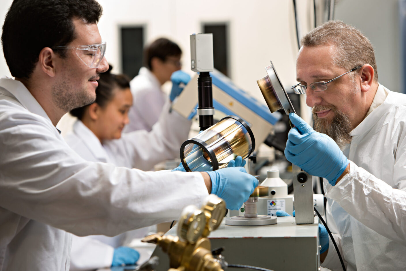 A team of researchers in goggles, gloves, and white coats, examining a component.