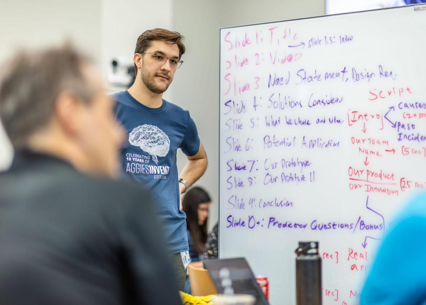 A man wearing a blue shirt that says "Aggies Invent," standing next to a large whiteboard filled with presentation notes.