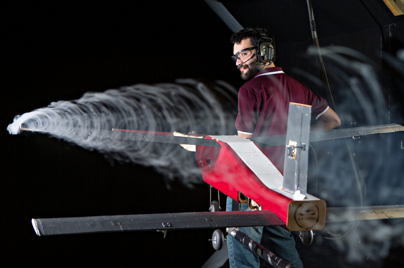 A person wearing safety gear stands beside a red and silver model aircraft emitting a trail of white smoke against a dark background.