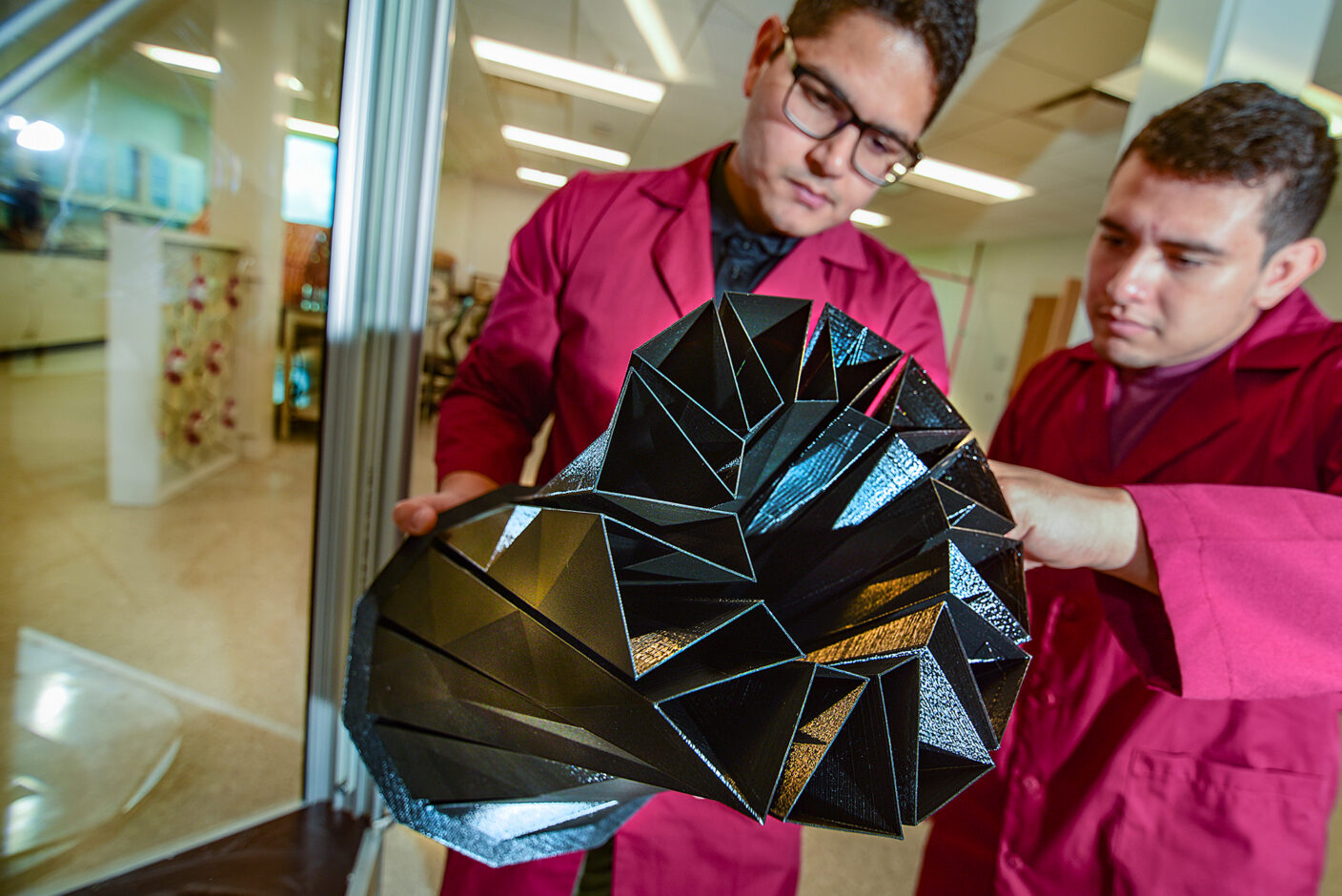 Two individuals in pink lab coats examine a complex, geometrically patterned black and metallic object together in a laboratory setting.