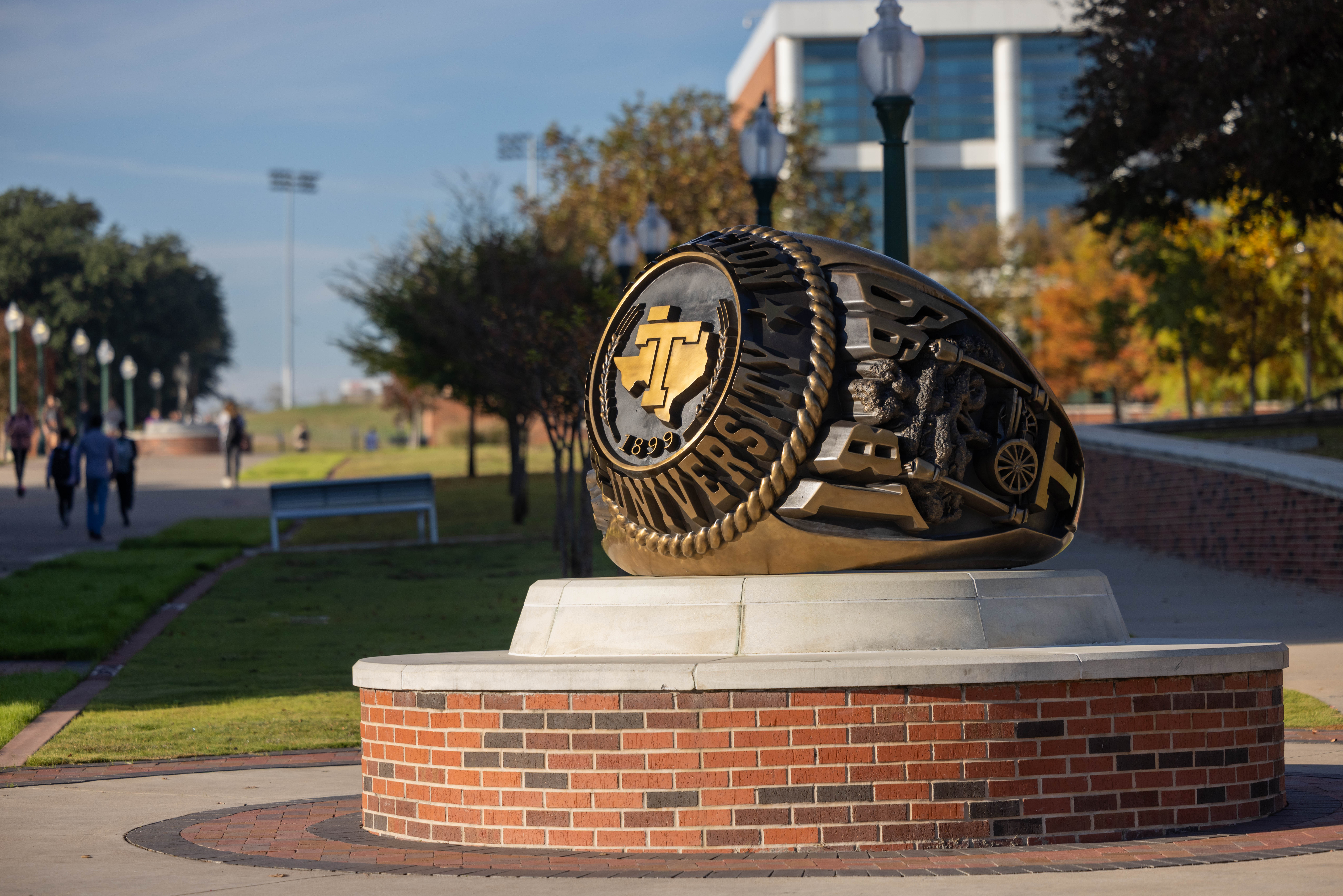 A large, ornate ring sculpture featuring the Texas A&M University logo and the year 1899 stands prominently on a brick pedestal in a campus setting.