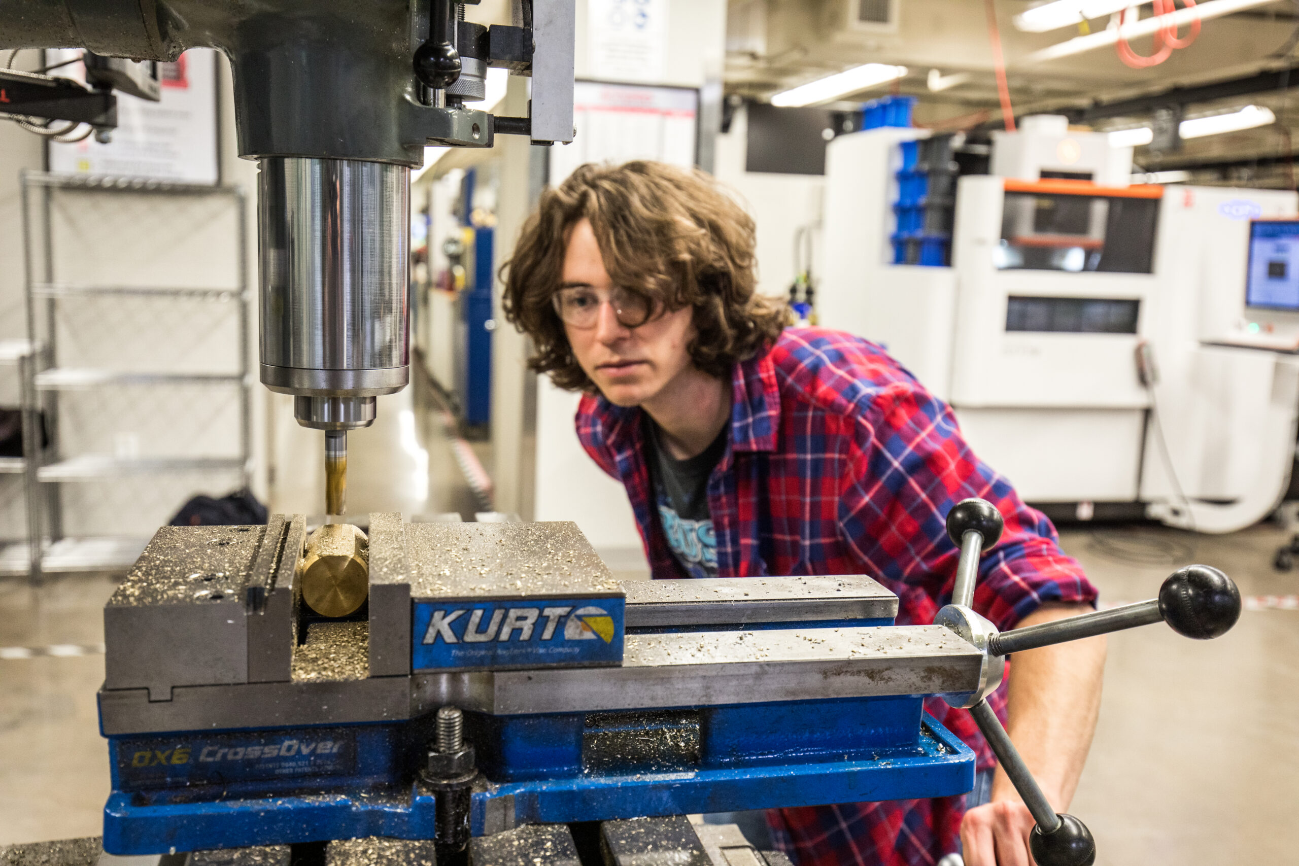 A person with curly hair and glasses operates a drill press in a workshop, carefully positioning a brass piece in a vise.