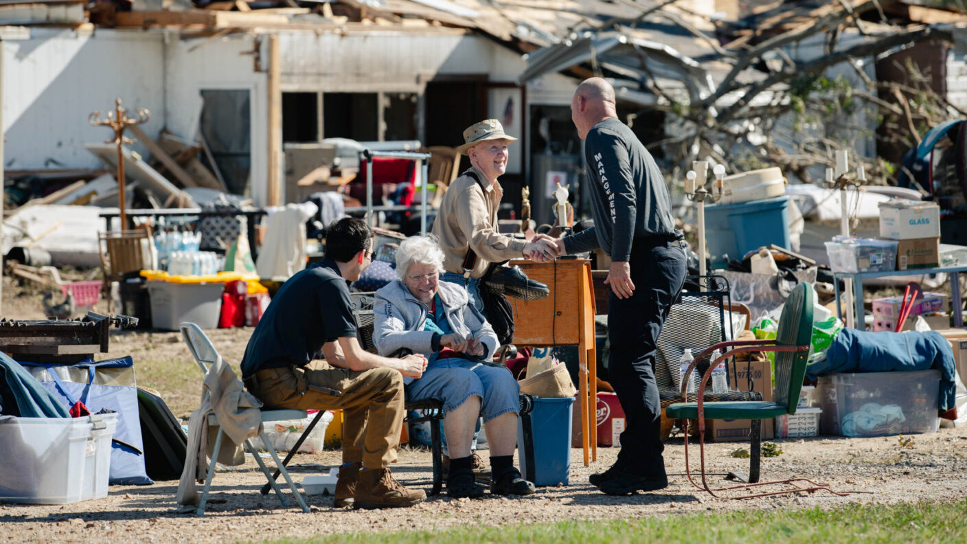 A group of people gather outdoors amidst debris and scattered belongings, with one individual shaking hands with another near a damaged building.