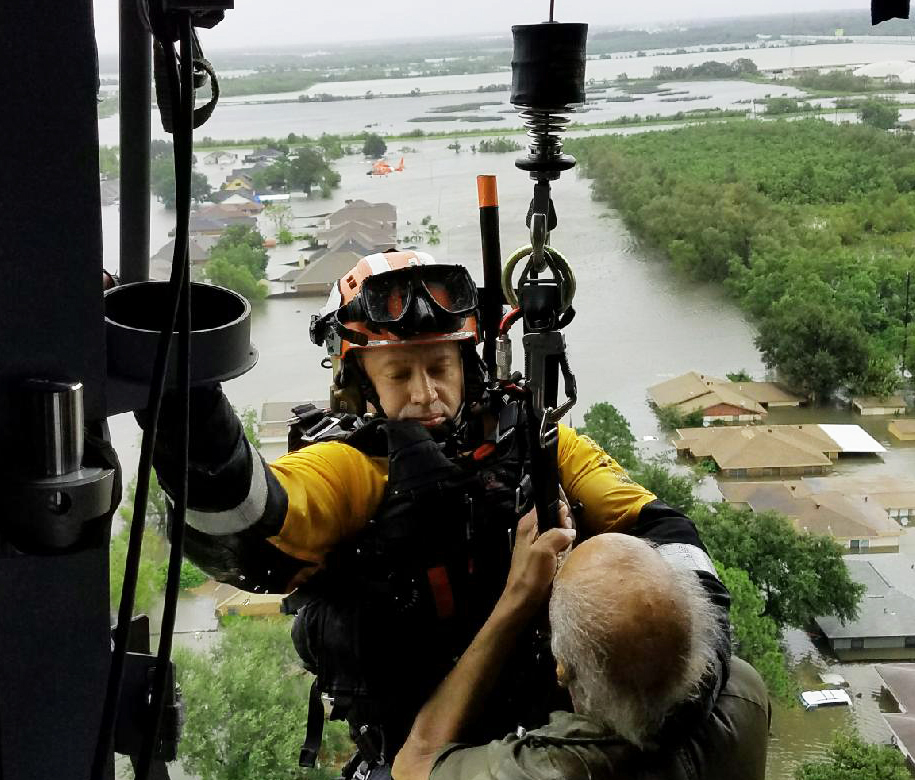 A rescue worker in a helicopter harnessed to a rescue line helps an elderly person during a flood rescue operation.