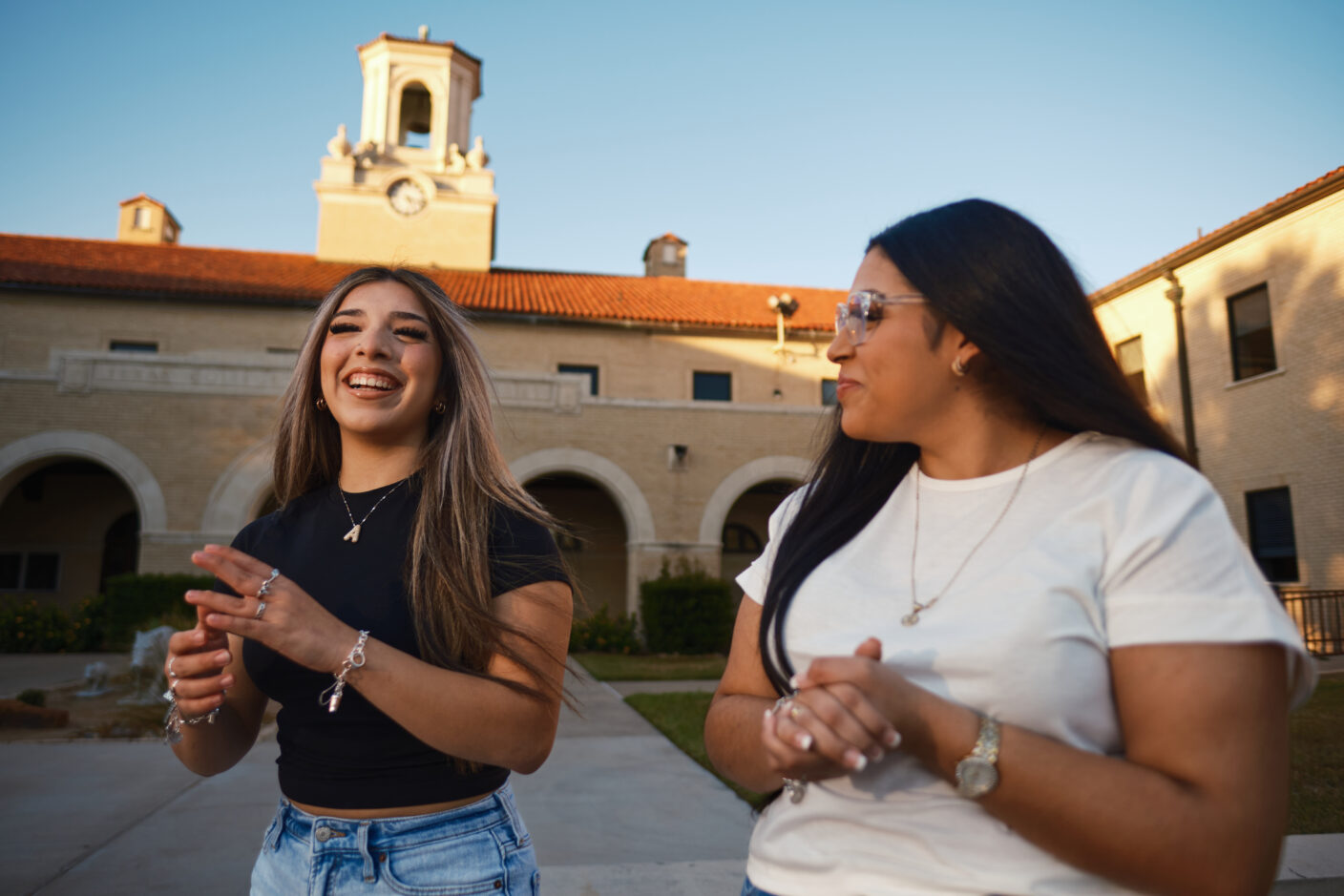Two individuals are engaged in a cheerful conversation outdoors, with a historic building featuring a clock tower in the background.