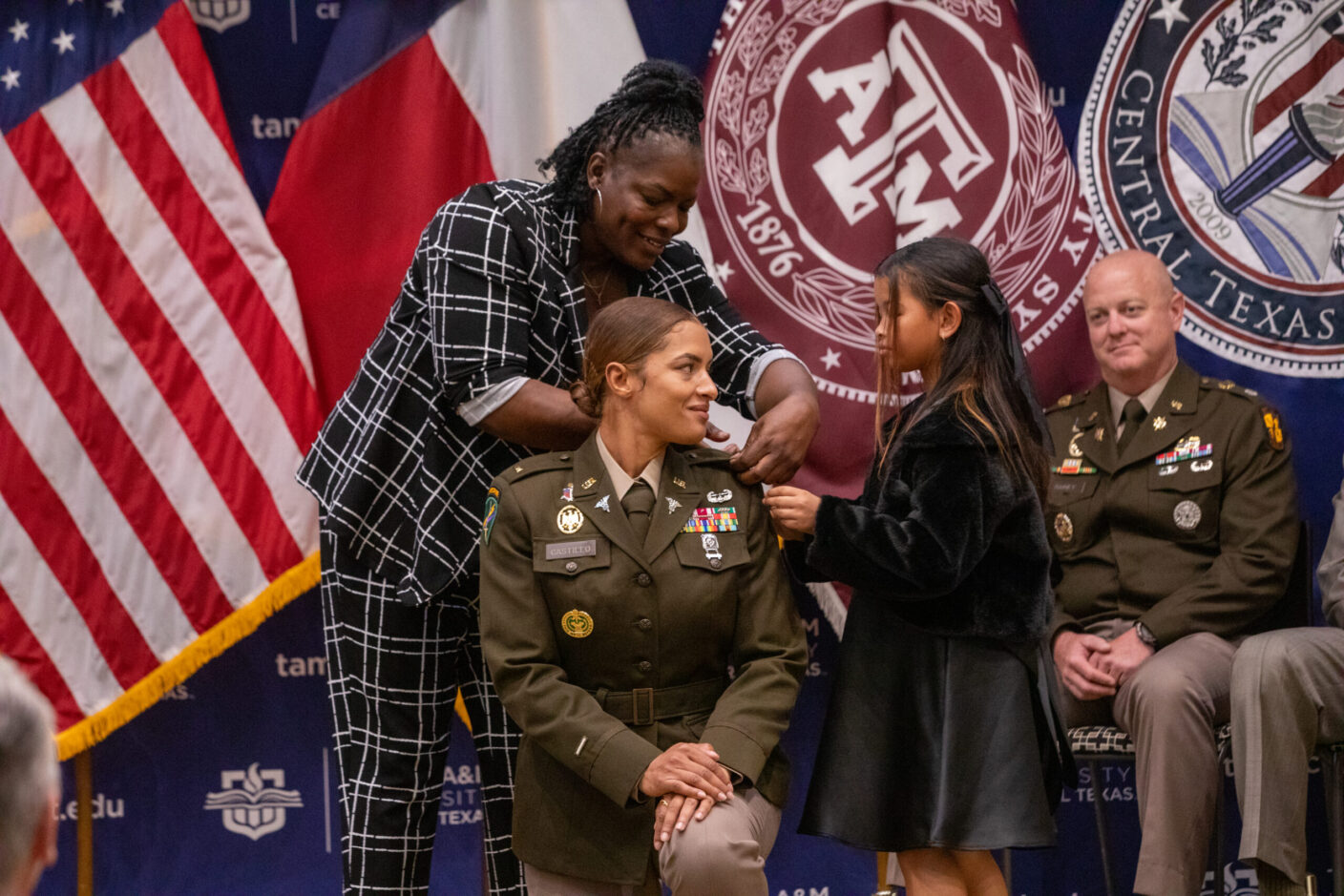 A person in military uniform is being pinned with a medal by a young girl, while another person assists, in front of flags and institutional banners.