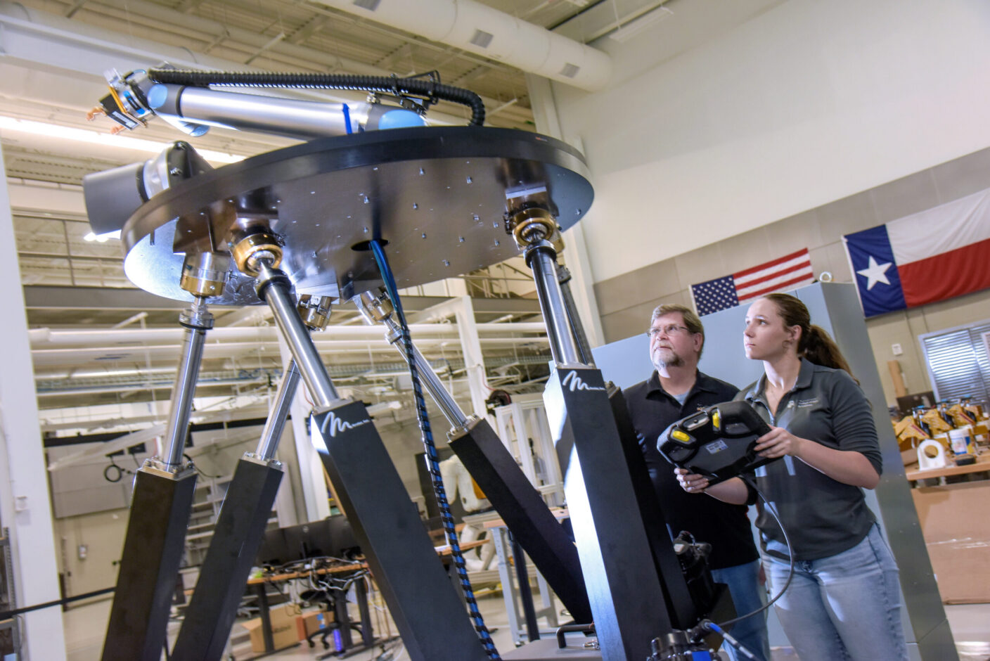 Two individuals operate a large robotic arm in a laboratory setting, with flags visible in the background.