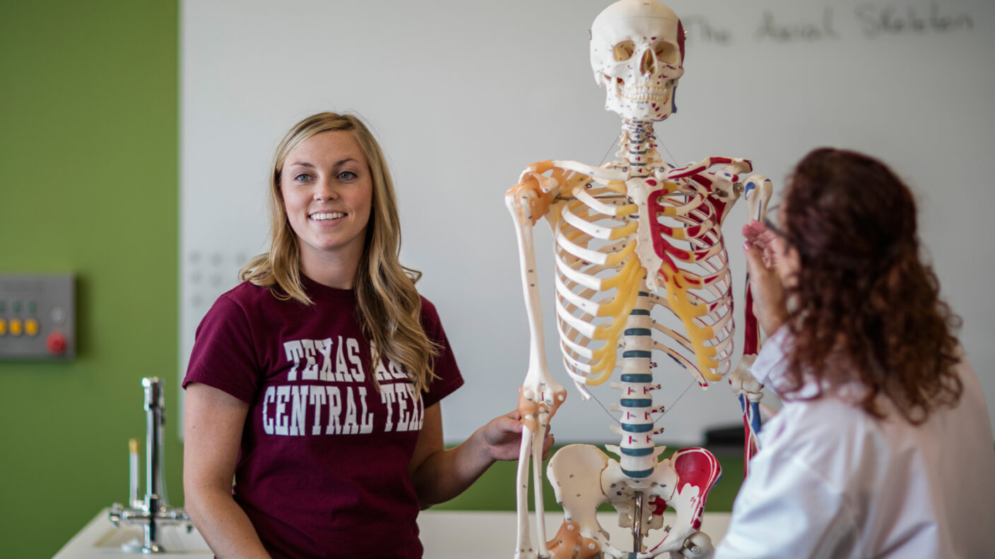 A student in a maroon shirt smiles while interacting with a detailed anatomical skeleton model in a classroom setting.
