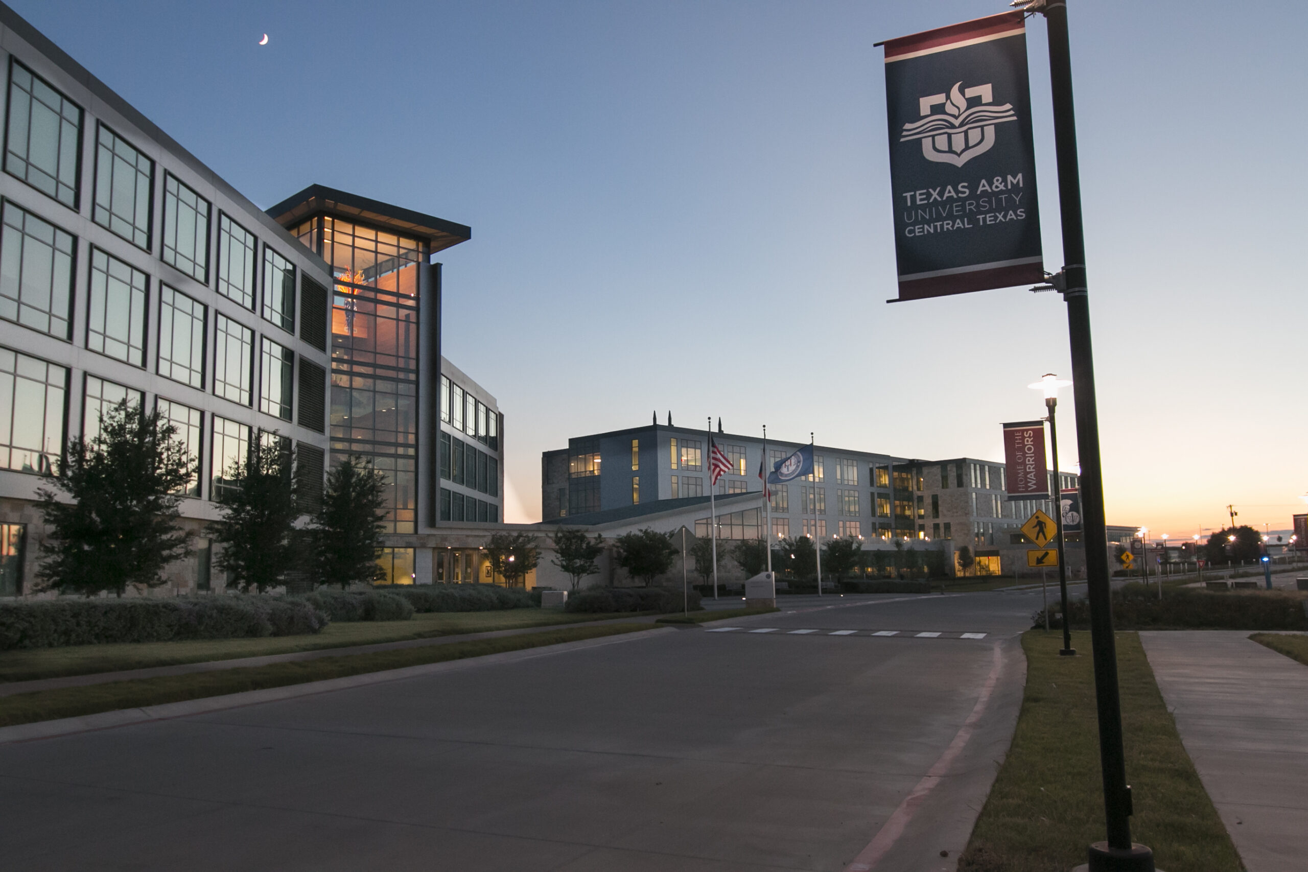 A banner for Texas A&M University-Central Texas hangs beside a paved road at dusk, with modern campus buildings and a crescent moon visible in the sky.