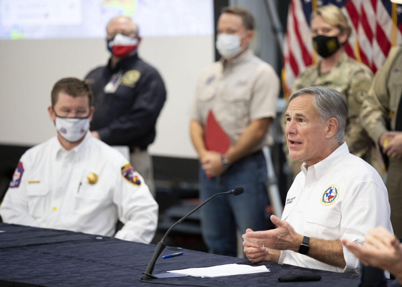 A person speaks at a table with a microphone during a press conference, while others in uniform and masks stand behind them.