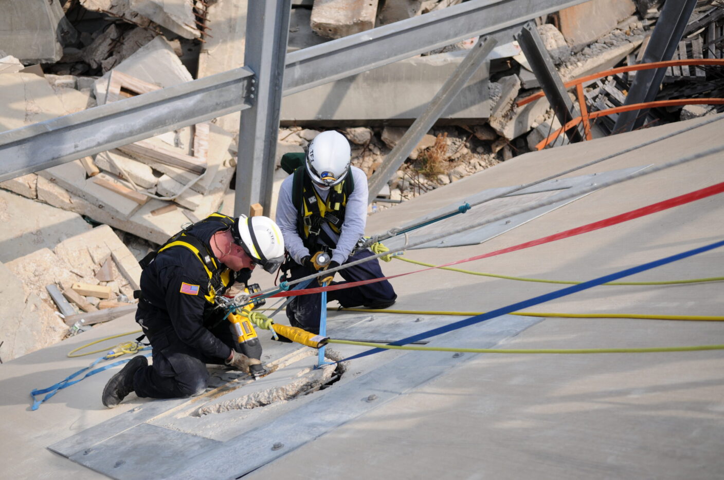 Two rescue workers in safety gear operate a tool on a damaged concrete surface amidst rubble and structural debris.