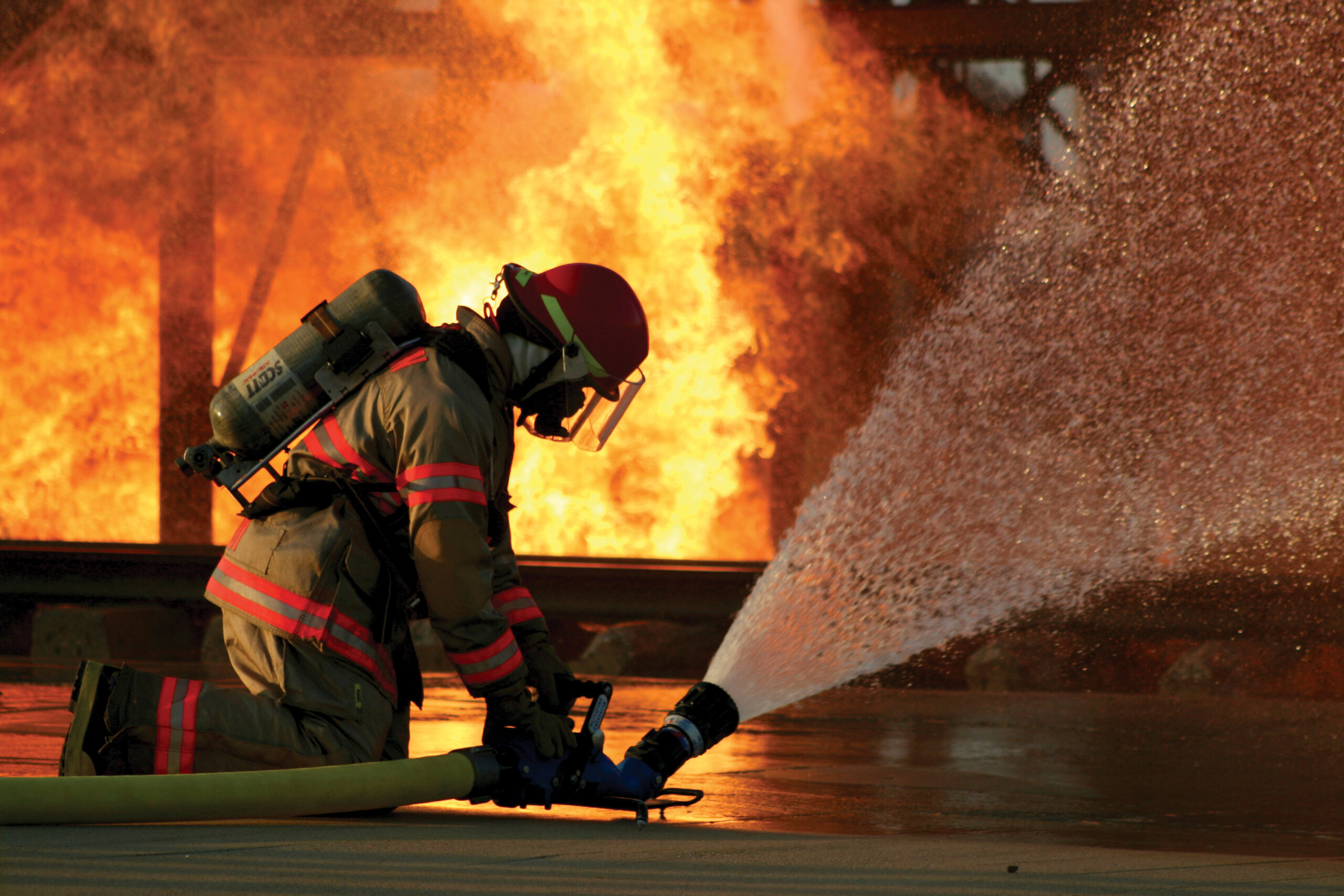 A firefighter kneels, spraying water from a hose to combat intense flames engulfing a structure.