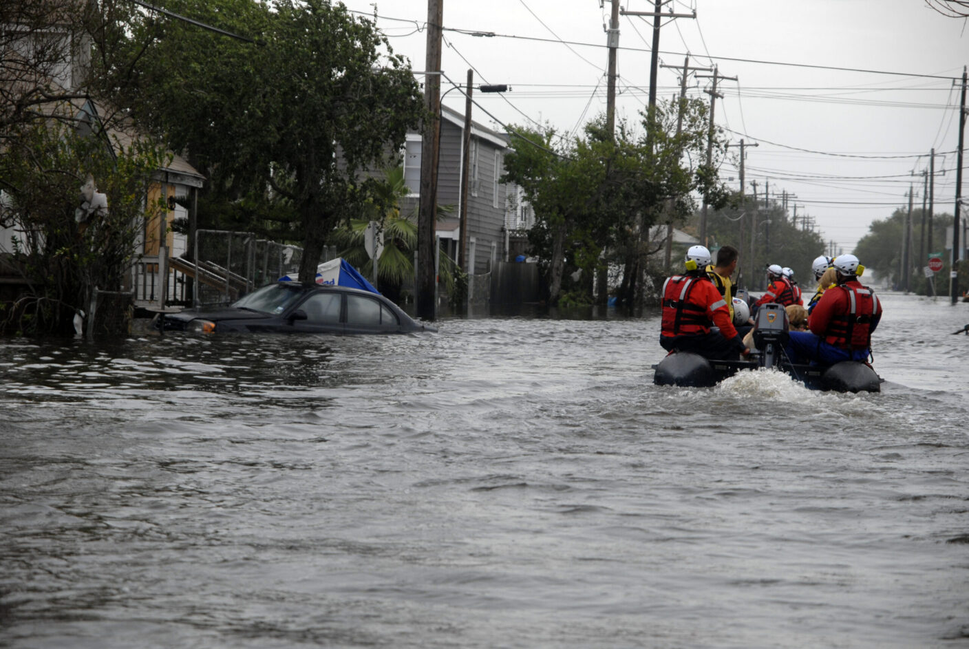 Rescue workers in a boat navigate through floodwaters past submerged vehicles and homes.