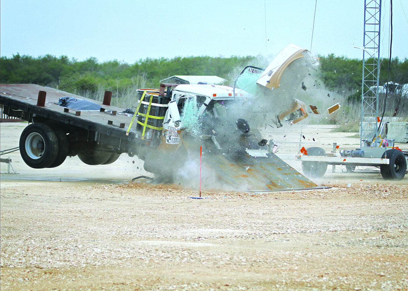 A white truck crashes into a large metal ramp, sending debris flying in a dusty explosion.