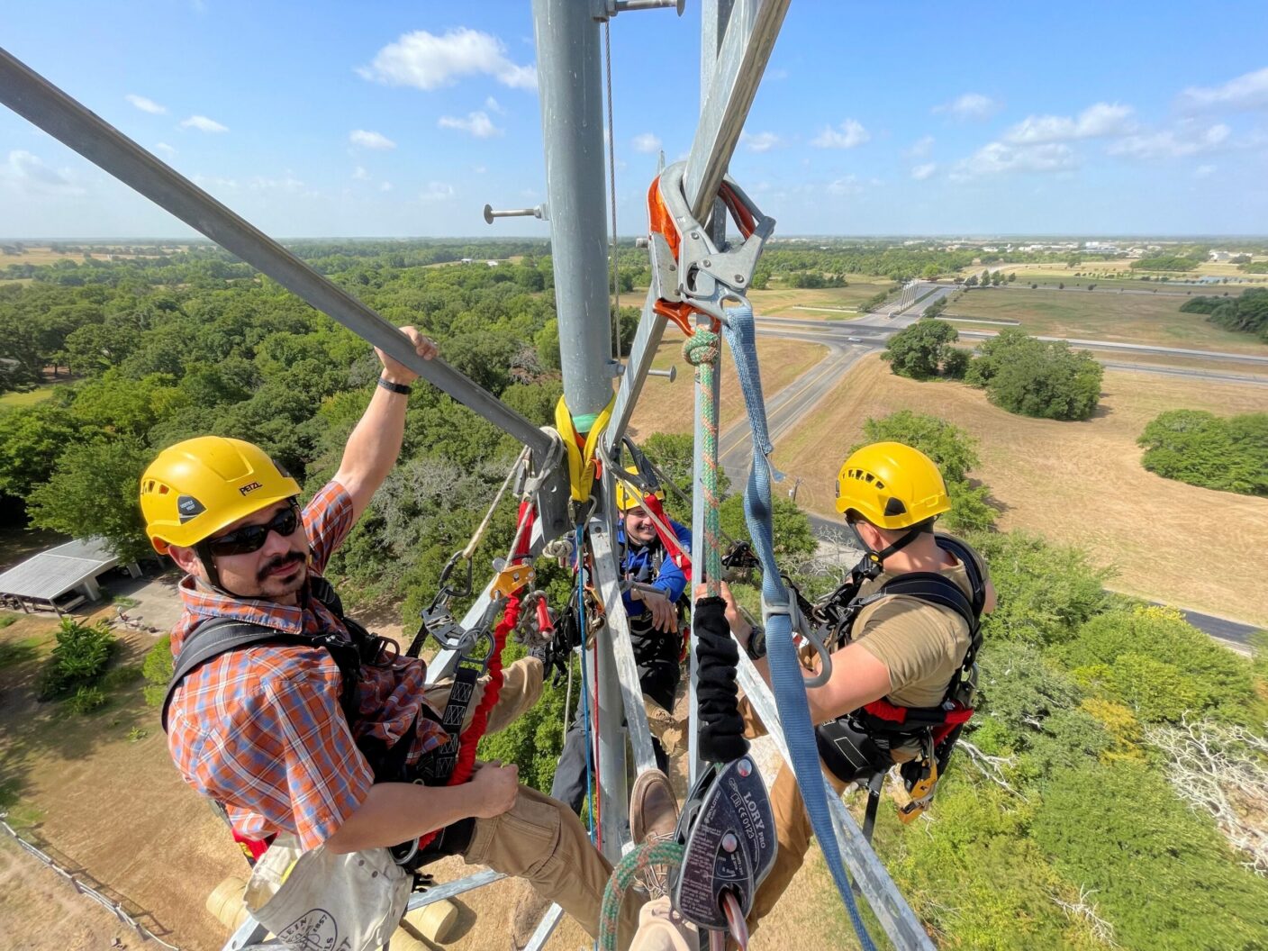 Three individuals in safety gear are working on a tall metal structure, suspended by ropes and harnesses, overlooking a landscape of trees and open fields.