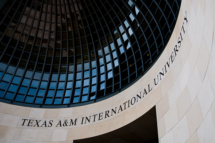 A curved stone facade with "TEXAS A&M INTERNATIONAL UNIVERSITY" engraved, beneath a large circular glass and metal structure.