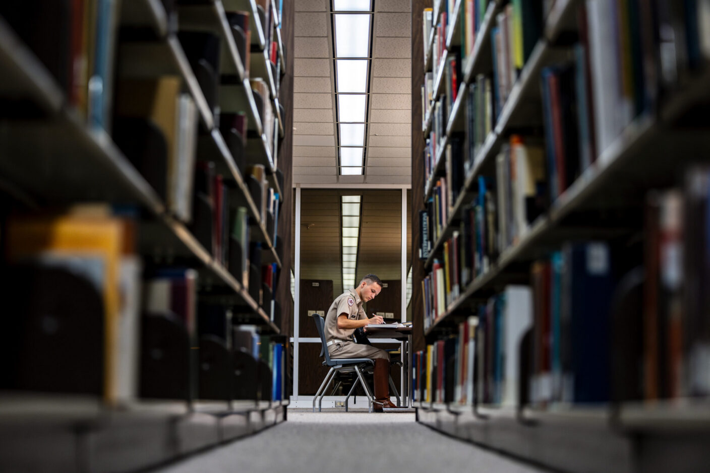 A person in uniform sits at a desk in a library aisle, surrounded by tall bookshelves, focused on writing.