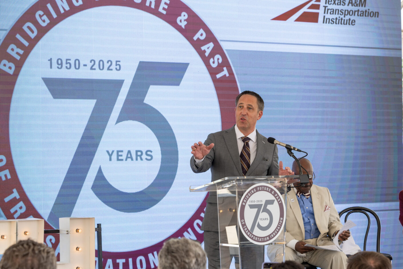 A person speaks at a podium during a 75th anniversary event for the Texas A&M Transportation Institute, with a large screen displaying "Bridging Future & Past 1950-2025."