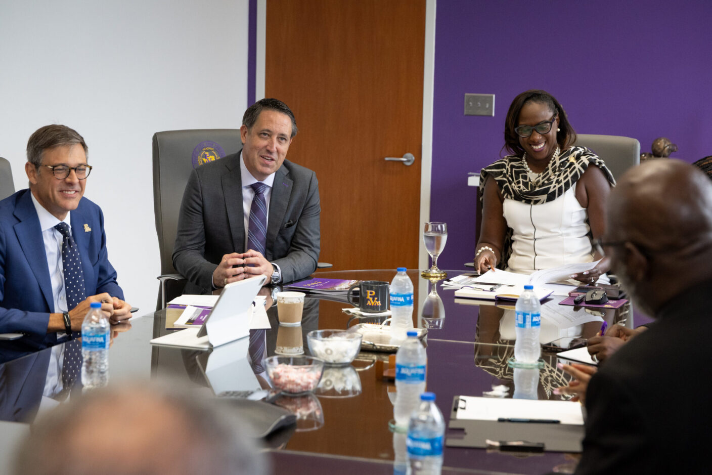 Several individuals in business attire are seated around a conference table, engaged in a meeting with smiles and active participation.
