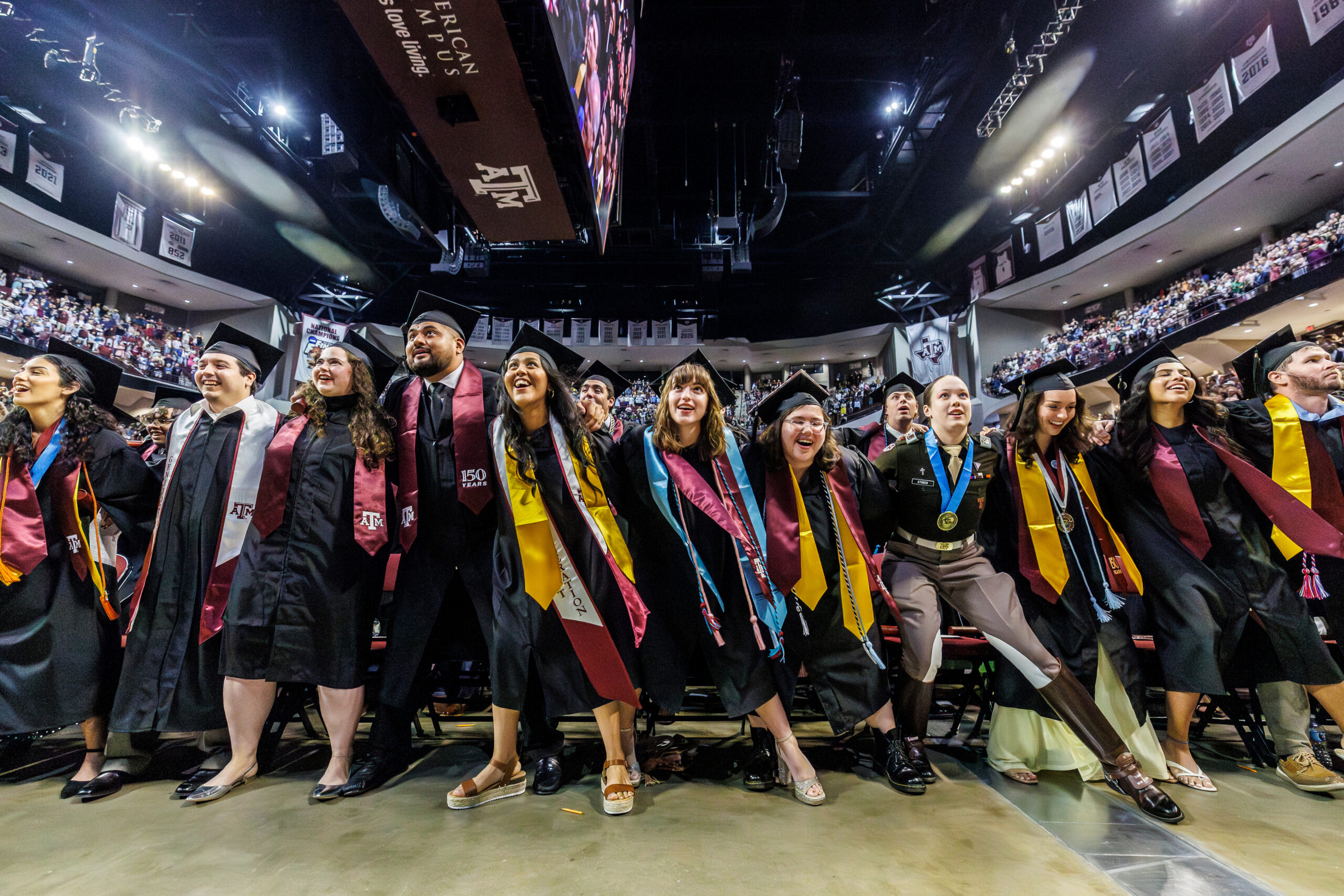 A group of graduates in caps and gowns with colorful stoles laugh and smile together on a stage during a large indoor ceremony.