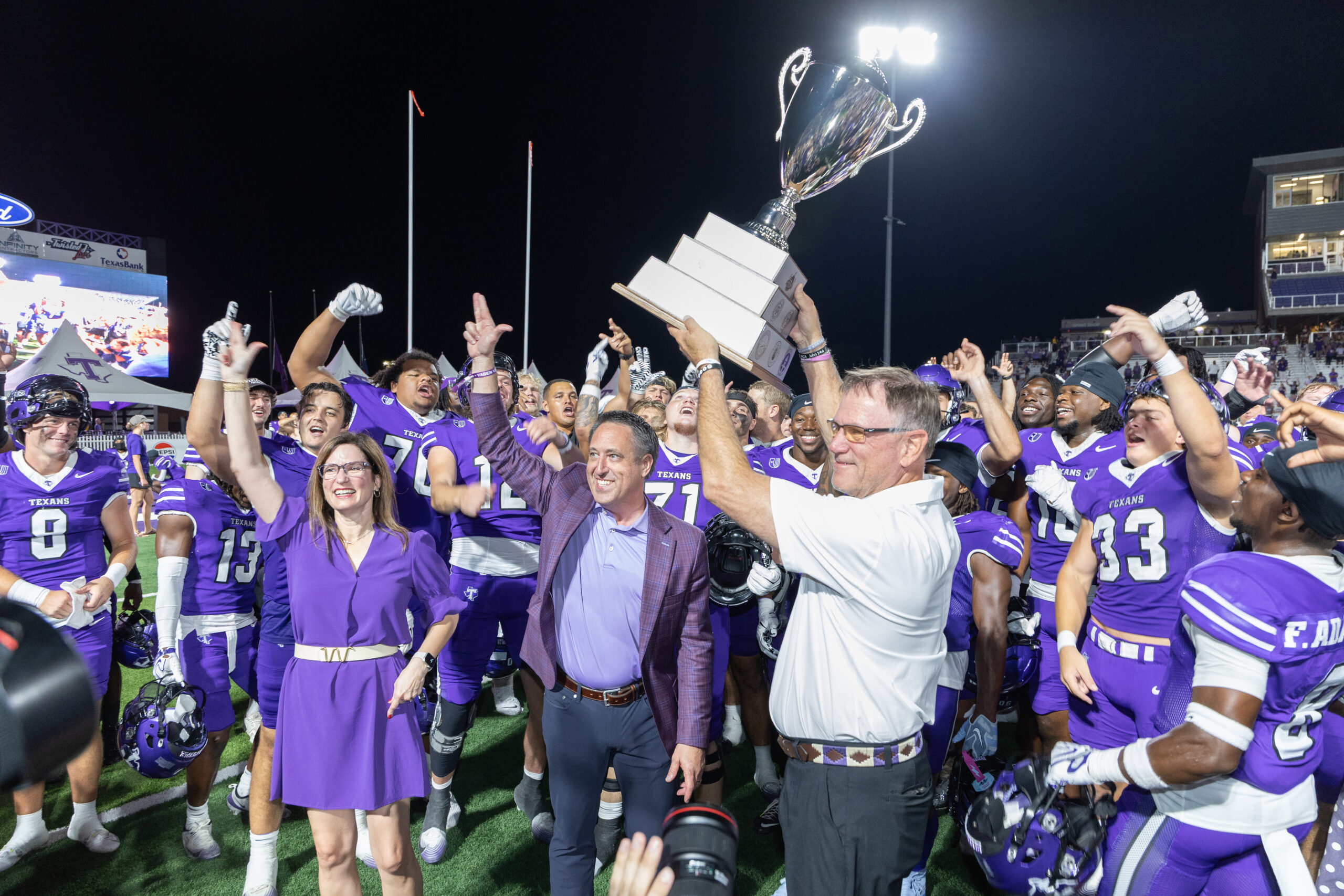 A group of football players in purple uniforms celebrate with a large trophy, surrounded by coaches and staff on a field at night.