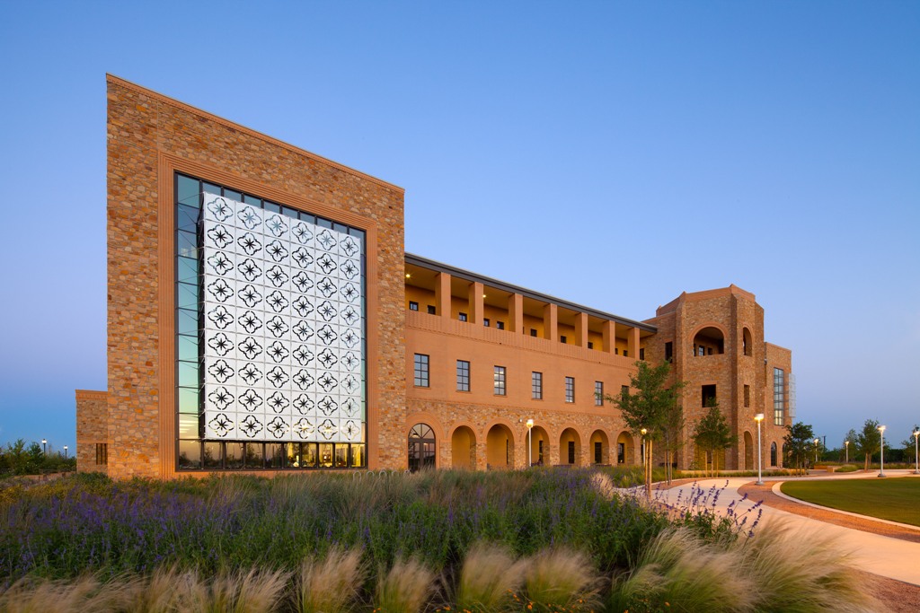 A modern building with a prominent stone facade and intricate glass paneling stands against a clear blue sky, surrounded by landscaped gardens and pathways.