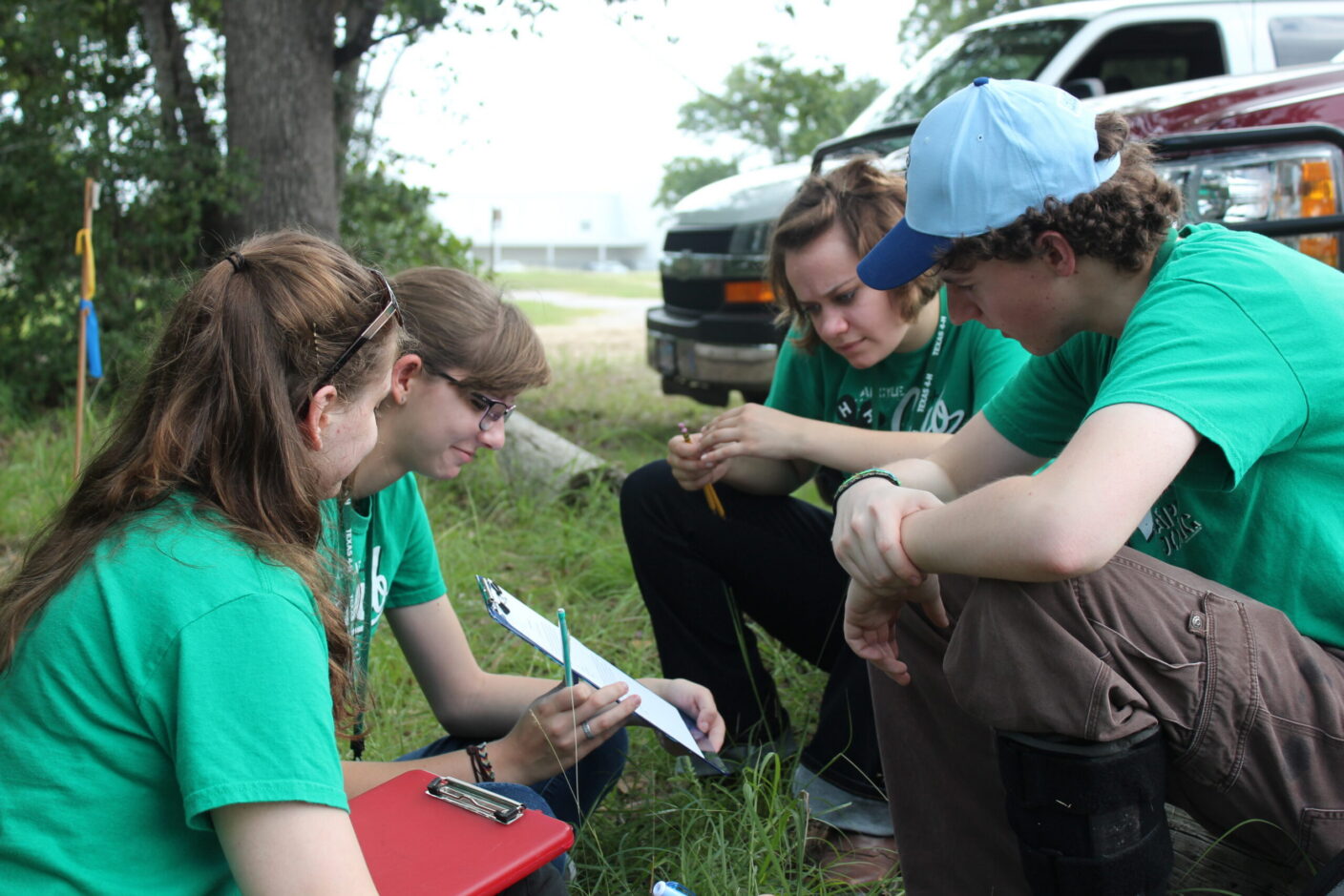 Four individuals in green shirts are crouched outdoors, examining something on the ground while one holds a clipboard and pen.