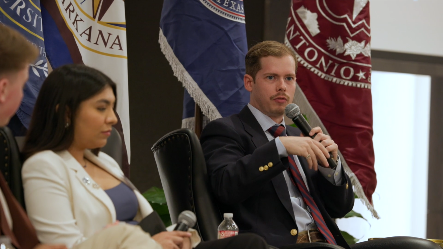 Two individuals participating in a panel discussion with university flags in the background.