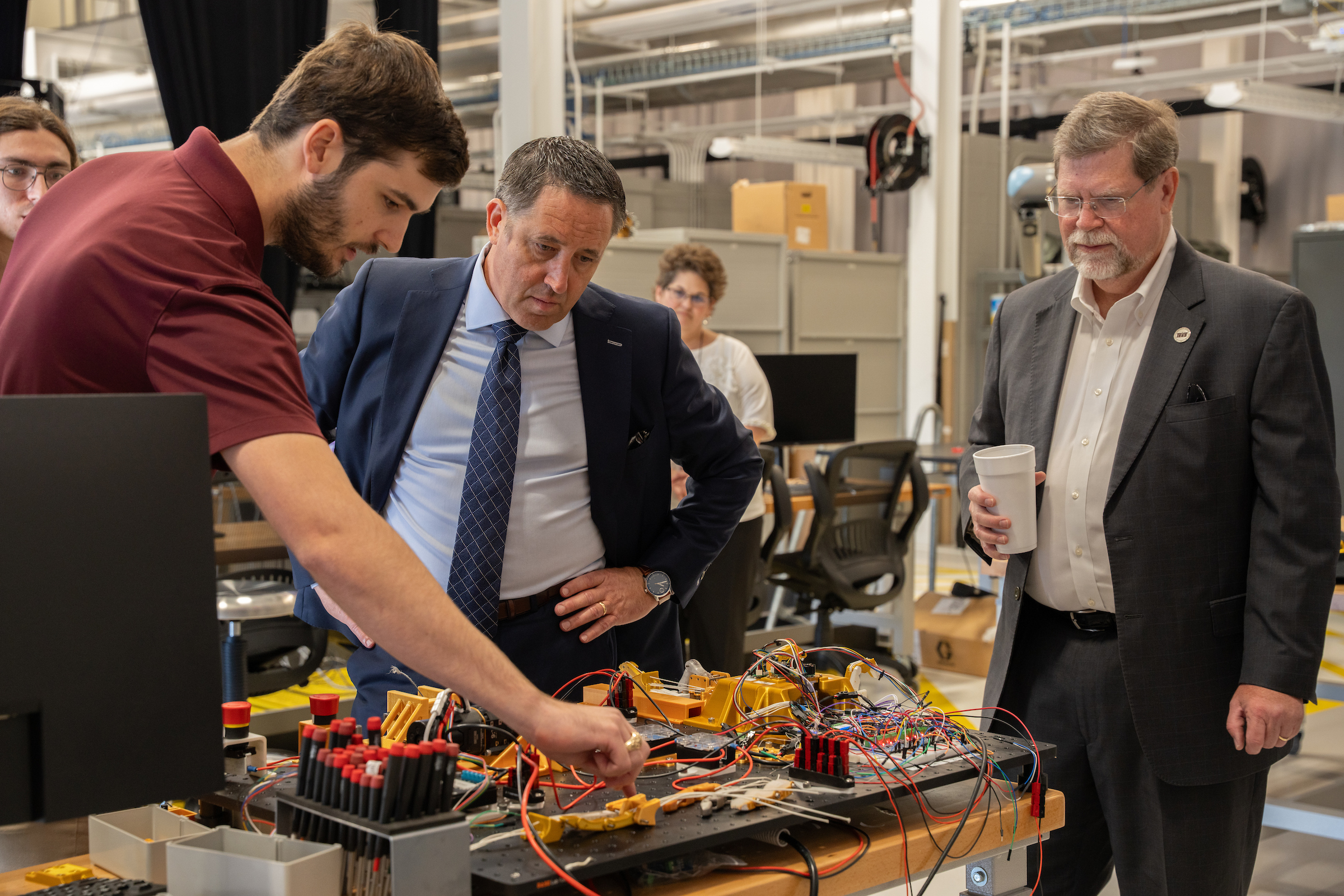 Three individuals examine a complex electronic circuit board in a workshop setting, with one person actively pointing to a component.