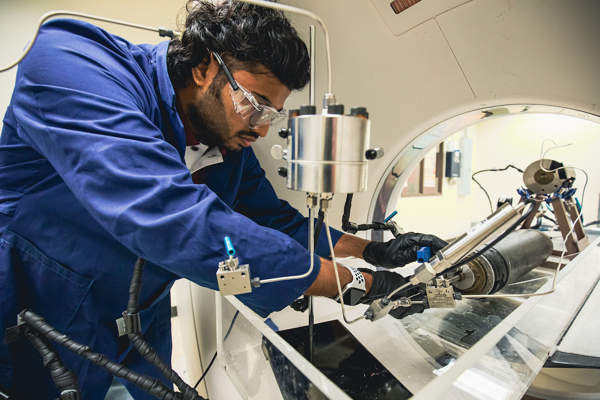 A technician in a blue lab coat and safety goggles carefully operates a complex piece of scientific equipment.