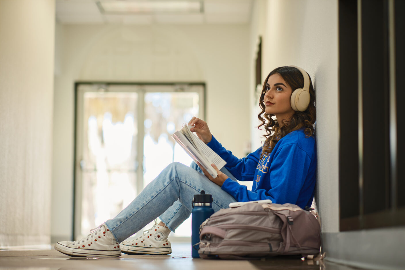 A person sits against a wall in a hallway, wearing headphones and reading a book while a water bottle and backpack rest nearby.