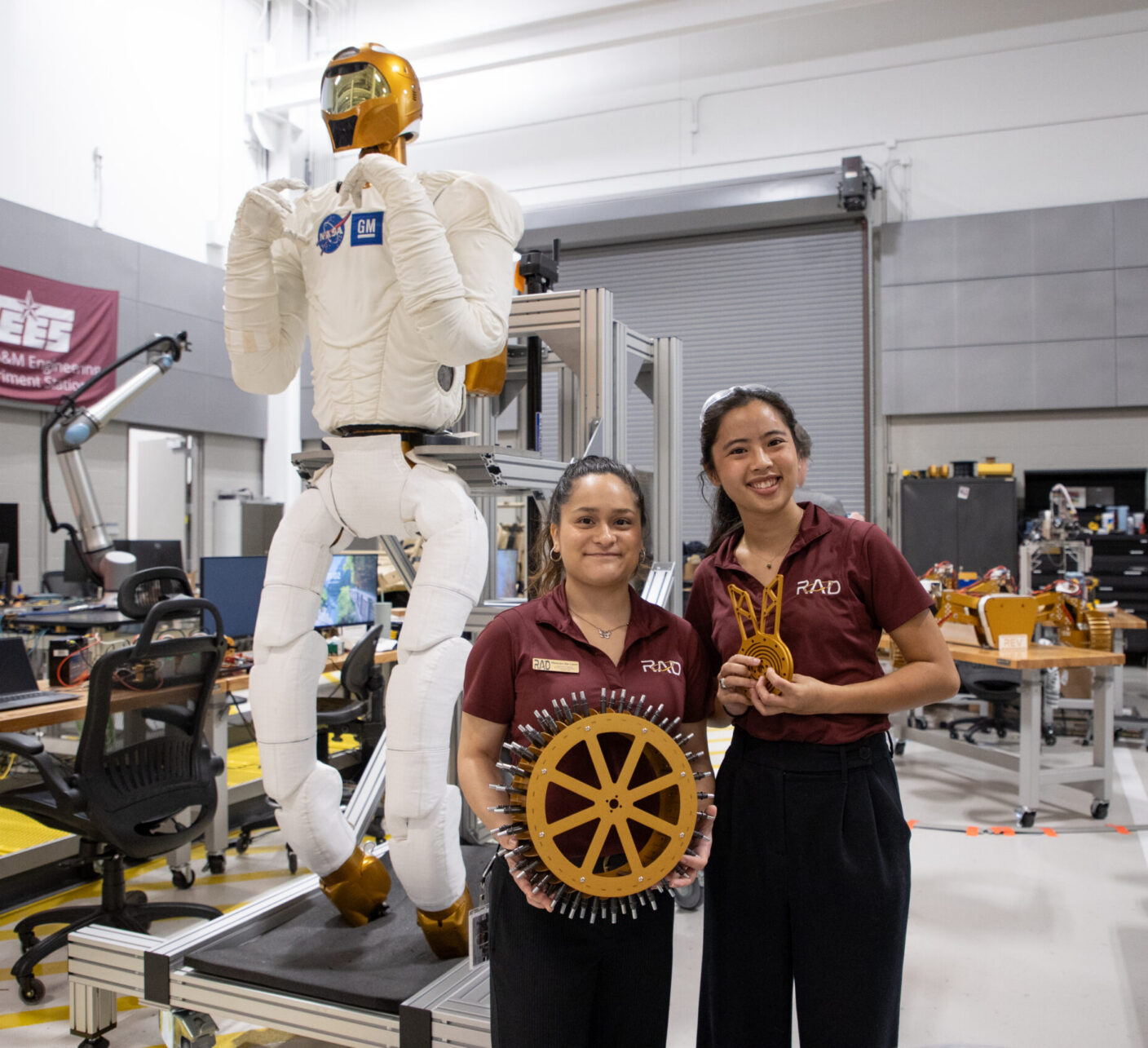 Two individuals stand in a workshop next to a tall humanoid robot, holding mechanical components.